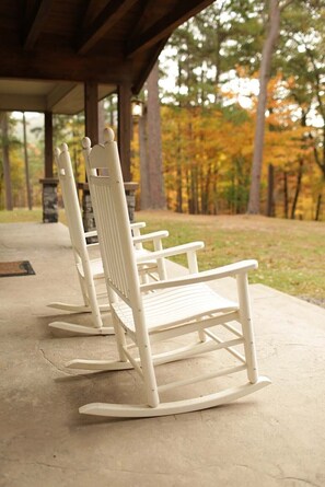 A row of white rocking chairs under a wooden shelter with a view of autumn trees.
