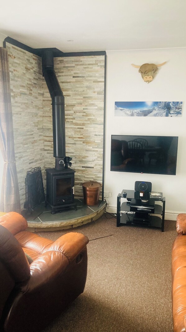 A living room with a stone wall, a black wood-burning stove, leather sofas, a flat-screen TV, and a mounted animal head decoration. - Number 2, The Morays. Two bedroom house (Blackford)