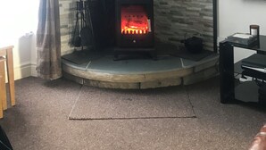 A corner room with a stone wall, a black wood-burning stove, a television mounted on the wall, and a window with curtains.