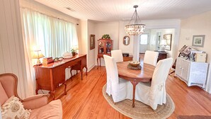 A dining room with a wooden table, white chairs, a chandelier, and a cabinet with glassware. - Vintage Cottage in Wanchese (Wanchese)