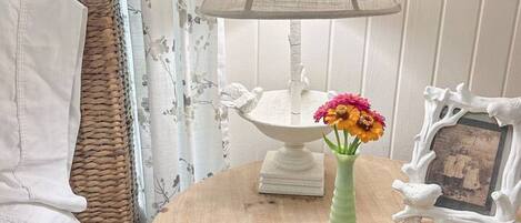 A round wooden table with a white lamp, a vase with flowers, and a picture frame.