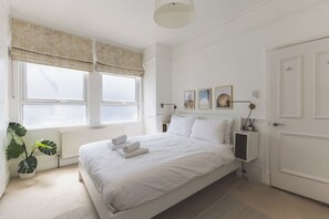 Een slaapkamer met een bed, een radiator, een raam met zonwering, een nachtkastje met lamp en een deur.