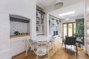 Een moderne keuken met witte bakstenen muren, een witte eettafel en stoelen. Er staan witte planken met planten en boeken.