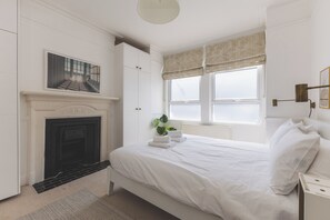 Ein Schlafzimmer mit Kamin, einem Bett mit weißen Leinen, einem Fenster mit Vorhängen und einem Gemälde über dem Kamin.
