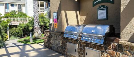 Ein Außenbereich mit Grillplatz, Steinmauern und Grillstationen, ein Schild mit Grillhinweisen sowie ein Blick auf ein Gebäude im Hintergrund.
