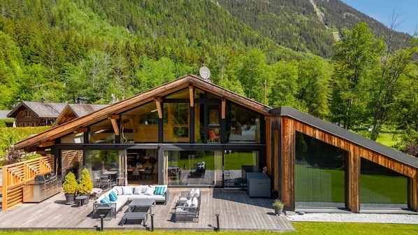 A modern wooden cabin with a deck, outdoor seating, and a mountain backdrop.