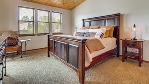 Quarto com teto de madeira, uma cama grande, mesa de cabeceira, cadeira e janela com persianas.
