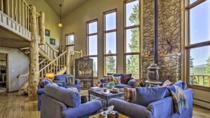 A spacious living room with a stone wall, large windows, and a rustic staircase. - Spacious Fairplay Retreat w/ Hot Tub & Fire Pit! (Fairplay)