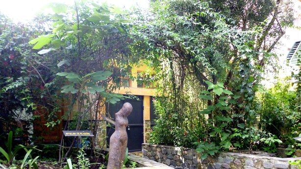 A stone pathway leading to a building with a yellow facade, surrounded by lush greenery and a stone wall.