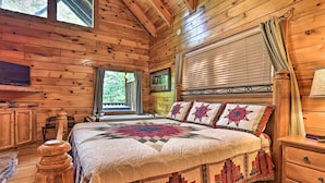 Ein Schlafzimmer in einer Holzhütte mit einem großen Bett, Holzwänden, einem Fenster mit Vorhängen, einem Nachttisch aus Holz und einem an der Wand befestigten Fernseher.