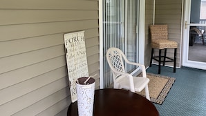 A porch with a sign listing porch rules, a wicker chair, a white plastic chair, and a table with a candle.