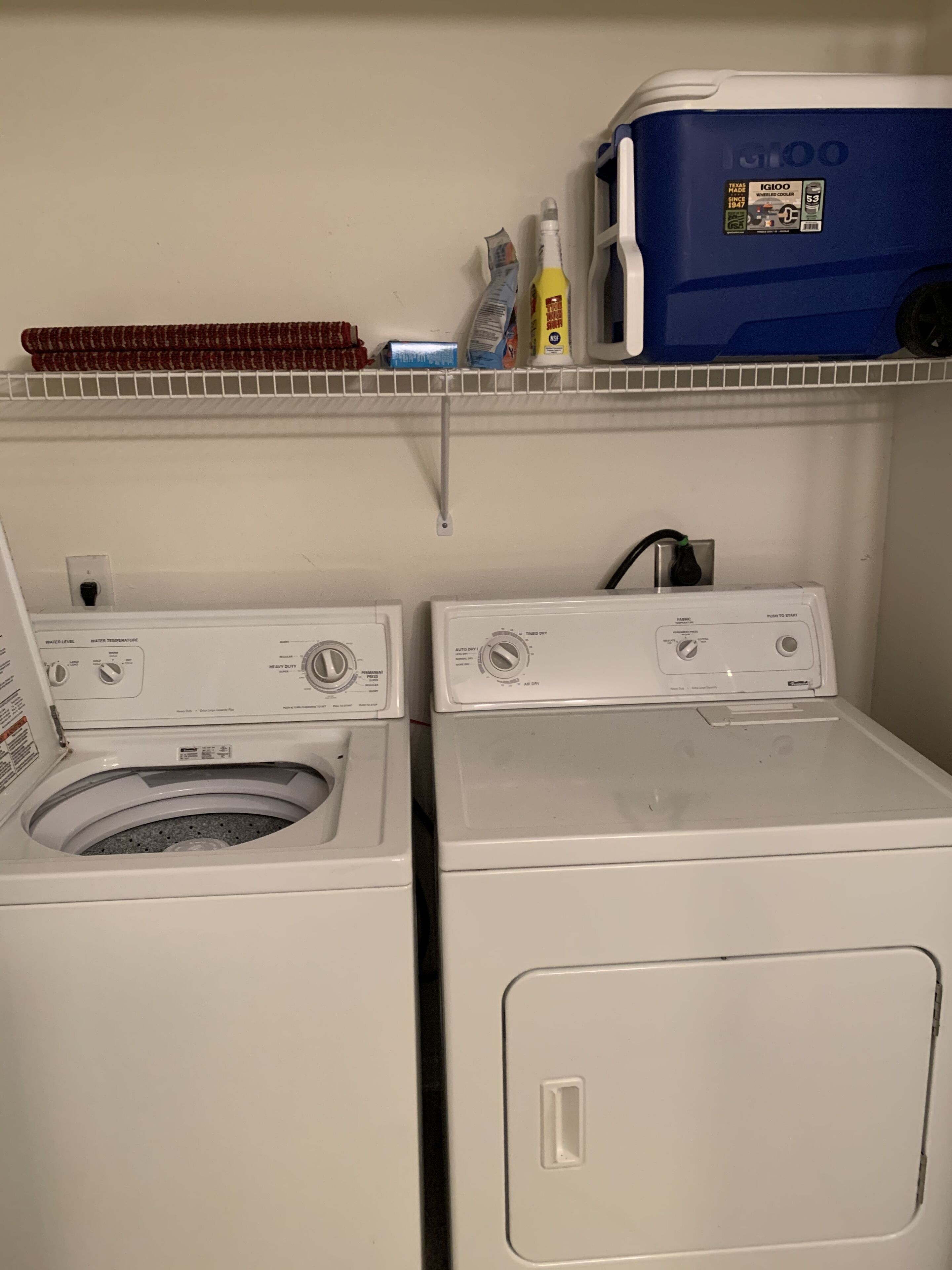 Un espace buanderie comprenant deux machines à laver et deux sèche-linge superposés, une étagère avec des produits de nettoyage et un récipient bleu portant l’étiquette « IG100 ».