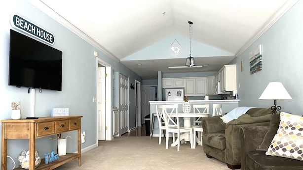 A living room with a TV, a wooden console table, and a couch. There is a kitchen area with a dining table and chairs.