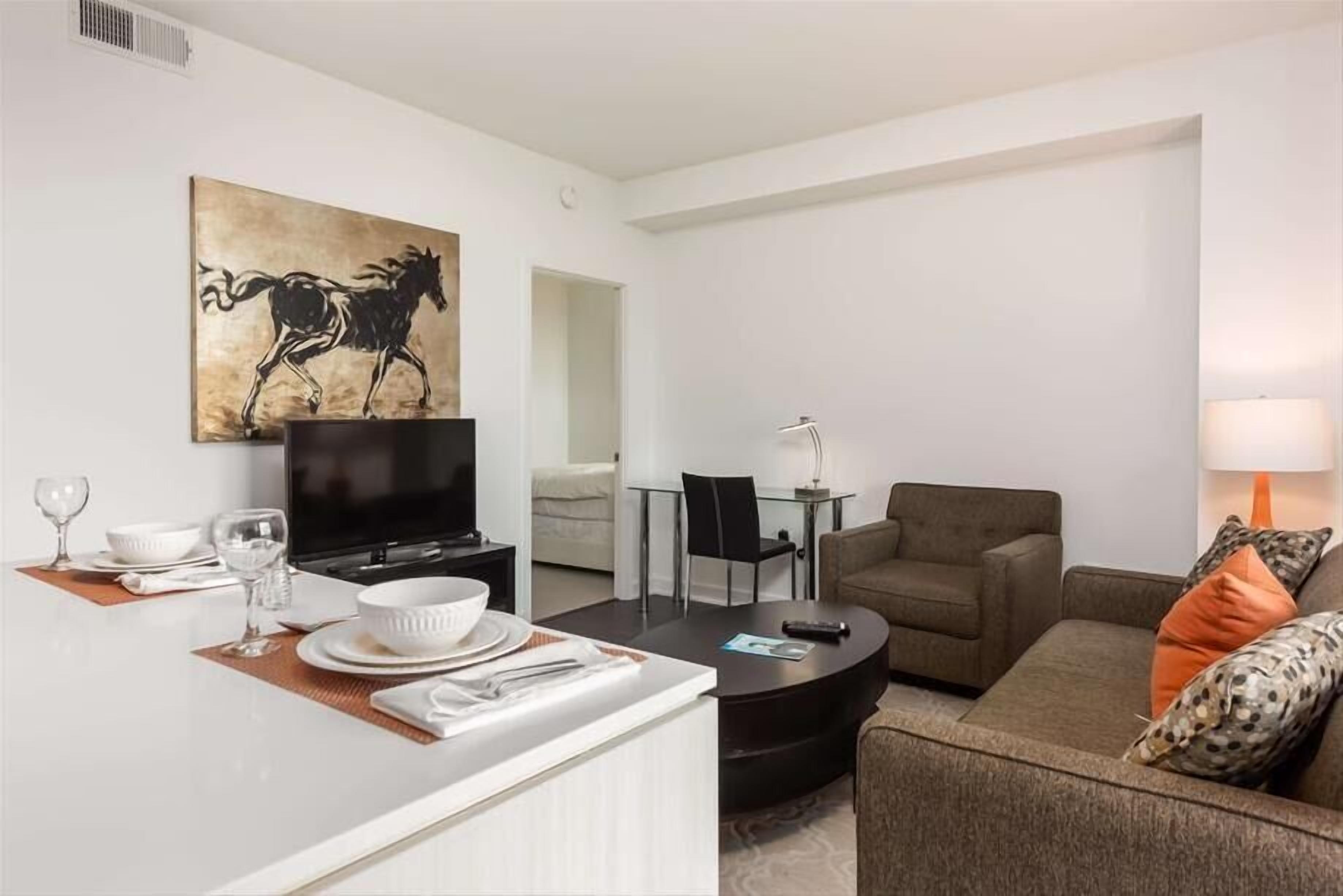 Ein modernes Wohnzimmer mit Flachbildfernseher, einem Essbereich mit weißem Tisch und Stühlen, einer Couch mit gemusterten Kissen und einem Bild eines Pferdes an der Wand.