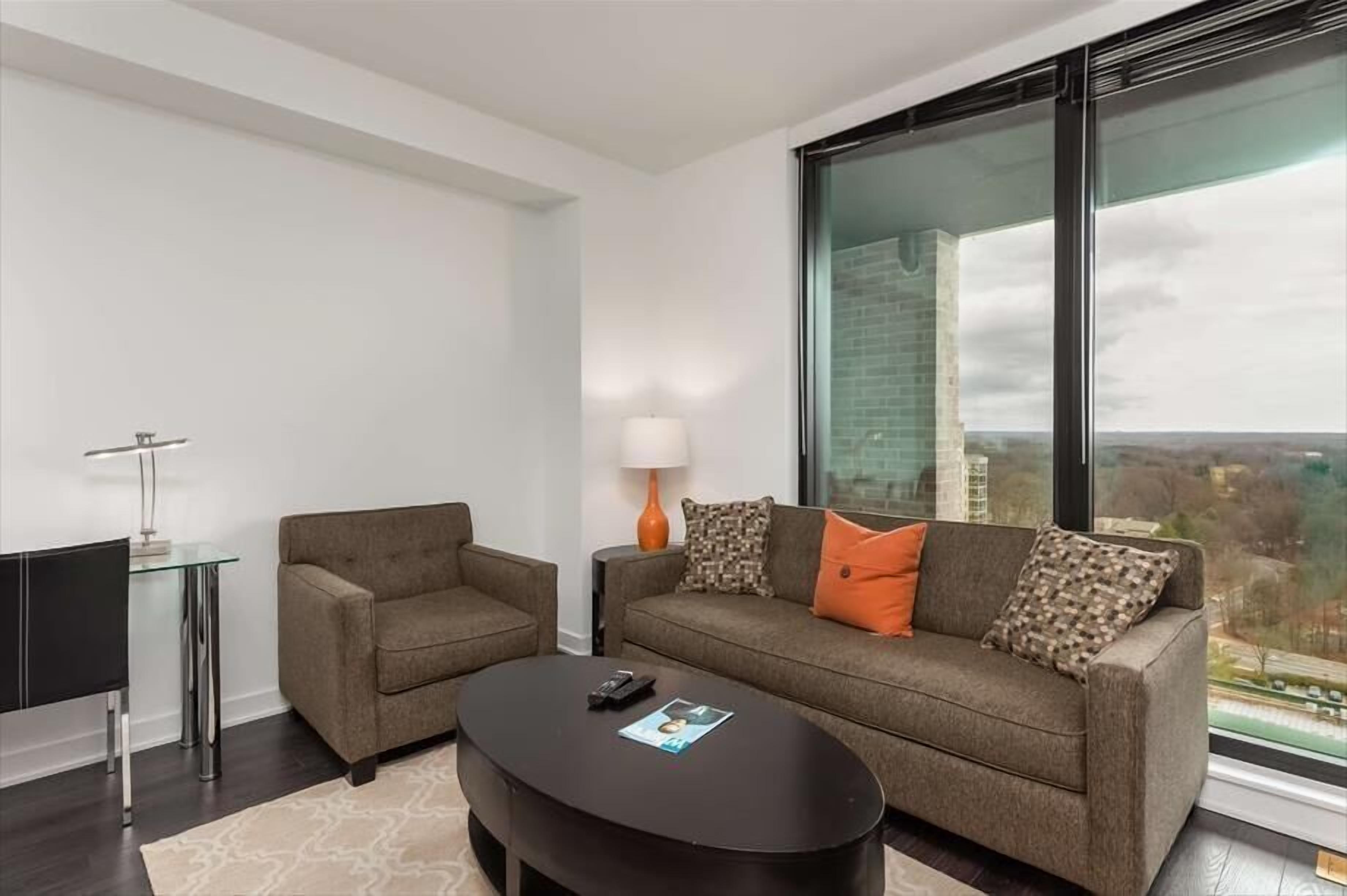 Una sala moderna con sofá, sillón y mesa de centro. Hay una mesa de vidrio con una lámpara y un televisor sobre un mueble bajo.