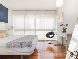 A modern hotel room with a large bed, a desk with a chair, a window with sheer curtains, and a potted plant on the desk.