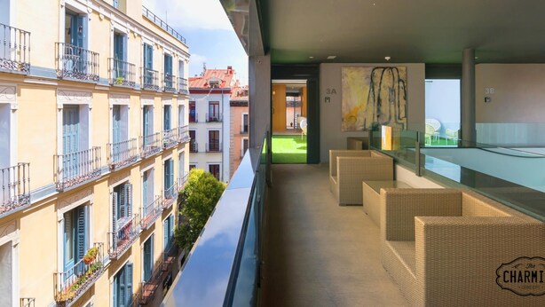 Un balcón moderno de hotel con mobiliario de mimbre, vista a edificios cercanos y un área verde al aire libre.