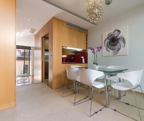 A modern dining area with a glass table and white chairs, a kitchen with wooden cabinets, and a large flower painting on the wall.