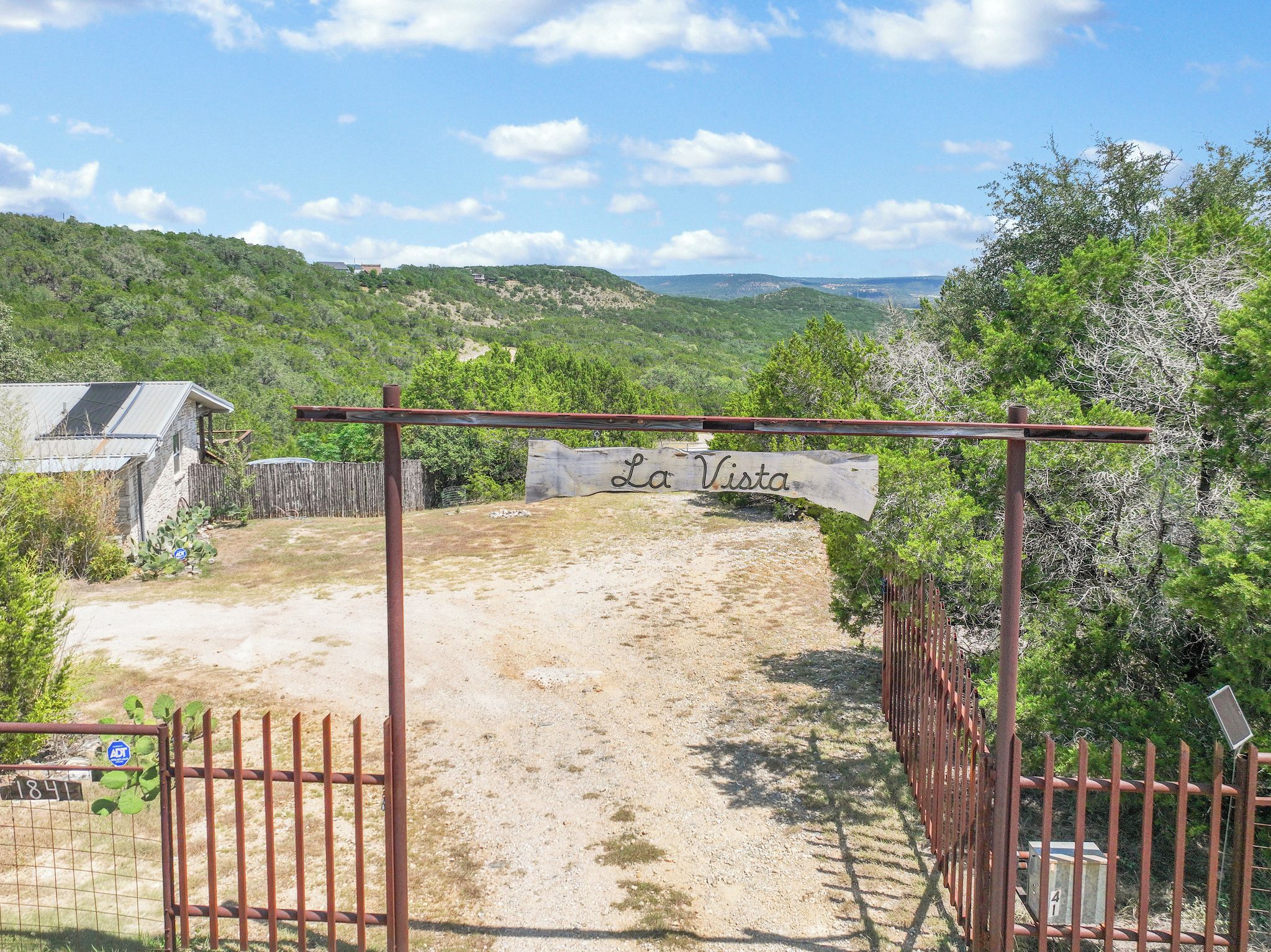 A gated entrance to a property named 'La Vista' with a dirt path leading inside, surrounded by greenery and hills in the background.