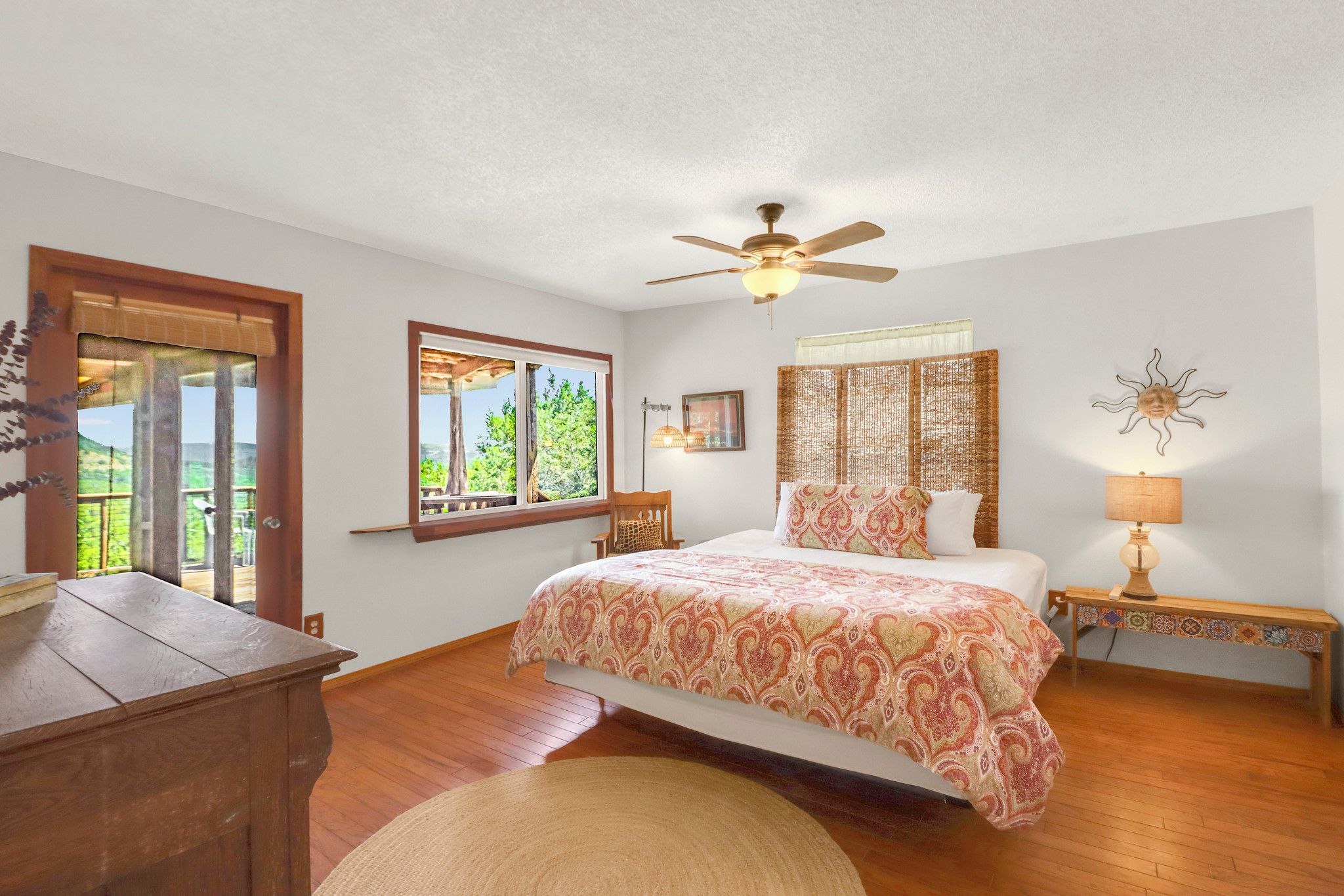 A bedroom with a bed, a ceiling fan, a nightstand with a lamp, a window with a view, and a wooden dresser.