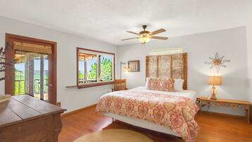 A bedroom with a bed, a ceiling fan, a nightstand with a lamp, a window with a view, and a wooden dresser.