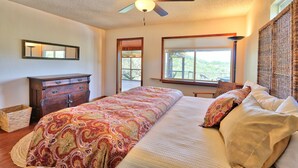 A bedroom with a large bed, a ceiling fan, a dresser, and a window with a view of greenery.