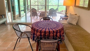 A cozy room with a round table, two wicker chairs, a sofa, and a sliding glass door leading to an outdoor patio with chairs and a lamp.