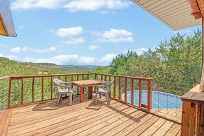 A wooden deck with a table and chairs, overlooking a pool and a mountainous landscape.