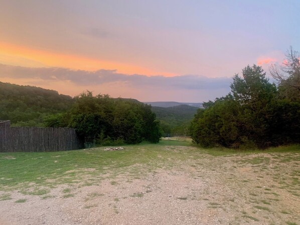 A rural landscape with a gravel path leading through a grassy area, surrounded by dense trees and a wooden fence.