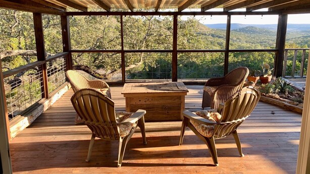 A covered outdoor seating area with wicker chairs and a wooden table, overlooking a mountainous landscape.