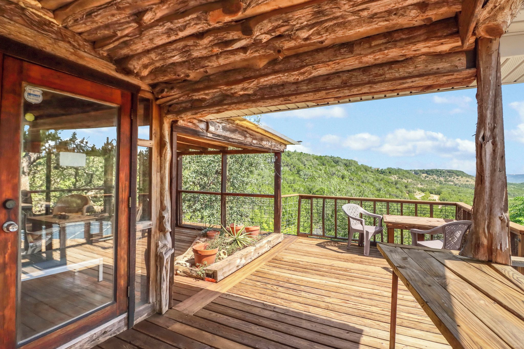 A wooden deck with a glass door leading to a room, a table and chairs, and potted plants.