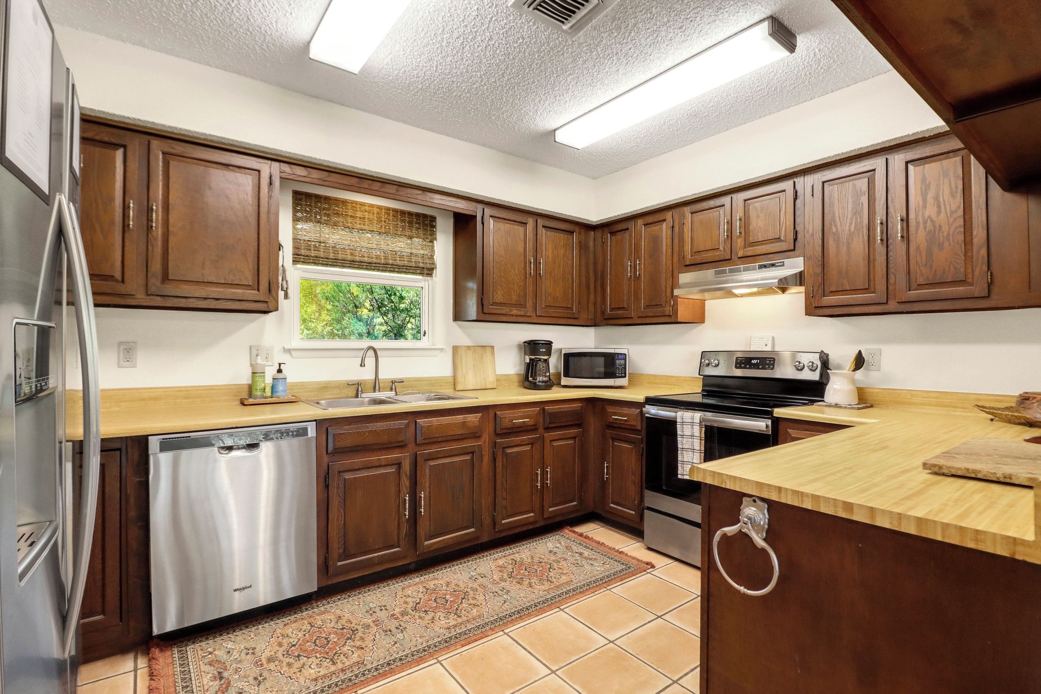 A kitchen with wooden cabinets, stainless steel appliances, a window with a view, and a tiled floor.
