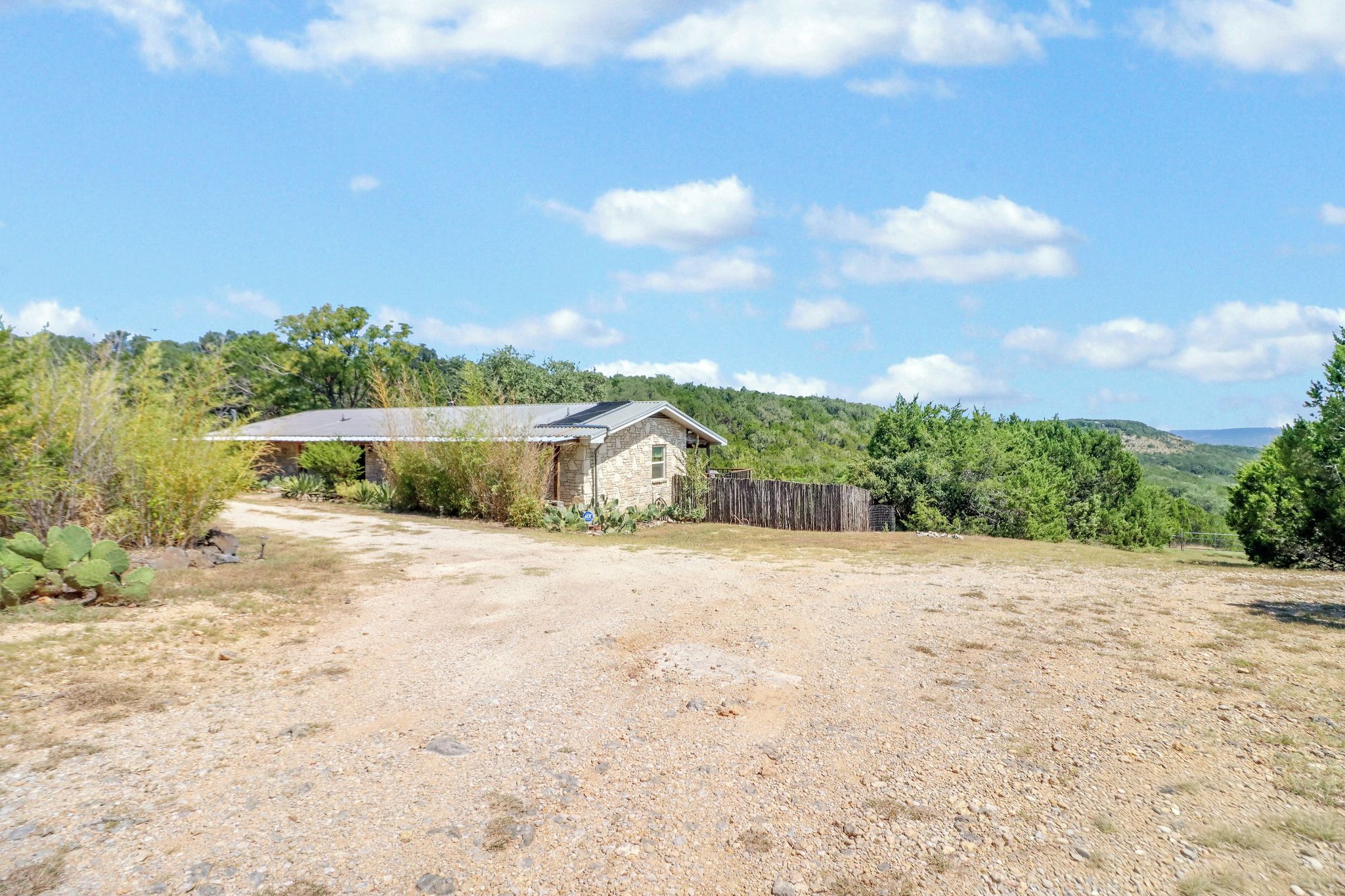 A rustic stone building with a metal roof, surrounded by a gravel driveway and a wooden fence.