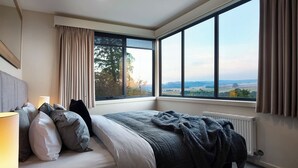 A bedroom with a large window, a bed with pillows, a lamp, and a radiator.