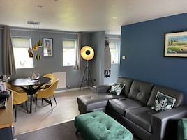 Uma sala de estar moderna com um sofá cinzento, um banquinho verde, uma mesa de jantar com cadeiras amarelas e um abajur.