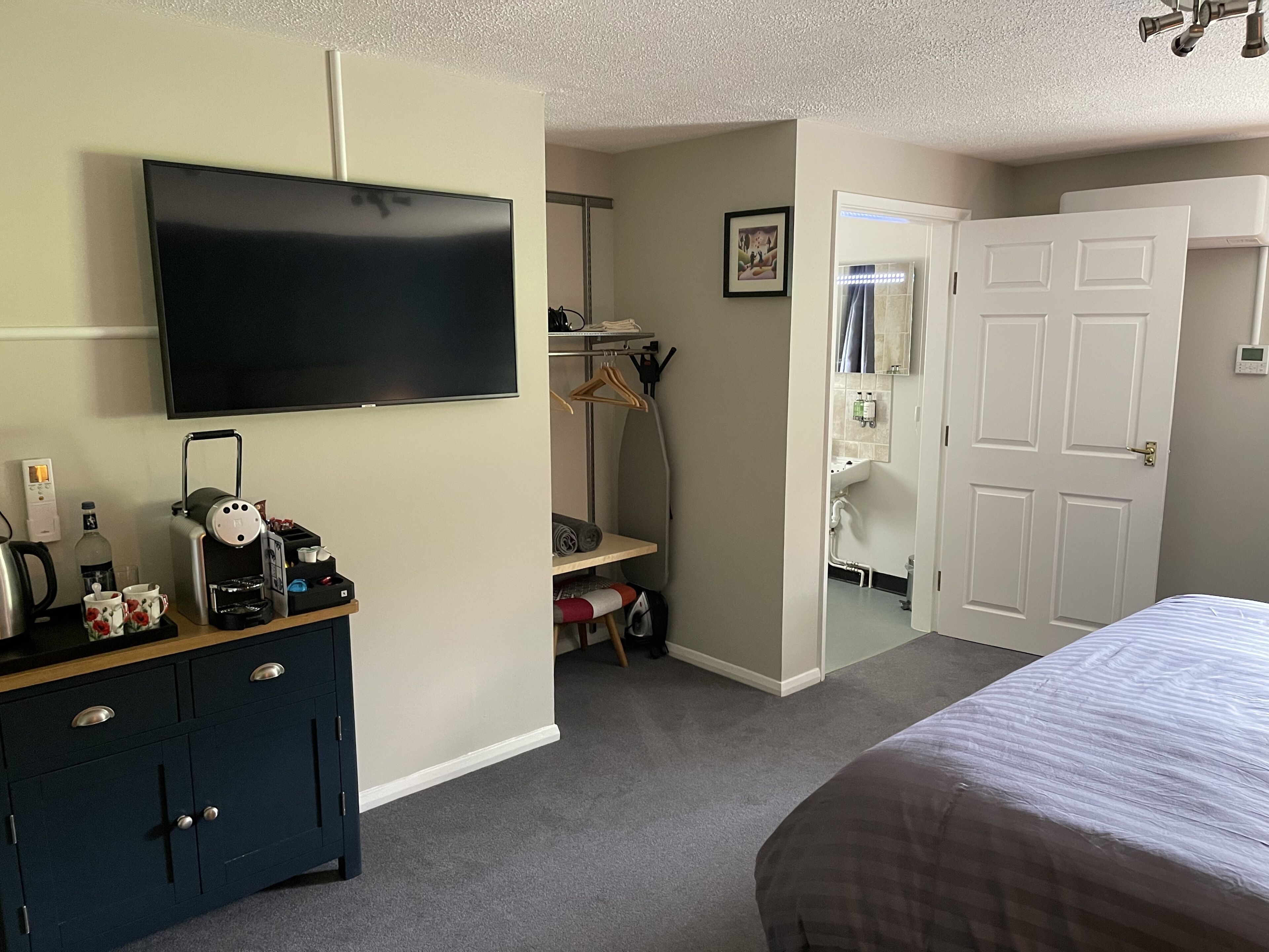 A hotel room with a flat-screen TV mounted on the wall, a dark blue dresser with drawers, a bed with a striped bedspread, and a door leading to a bathroom.