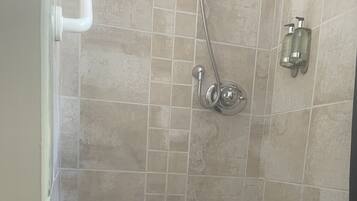 A shower with a handheld showerhead, a wall-mounted soap dispenser, and a drain in the floor.
