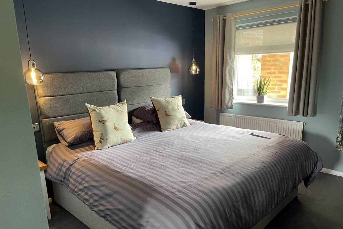 A bedroom with a bed, headboard, pillows, bedside lamps, and a window with curtains.