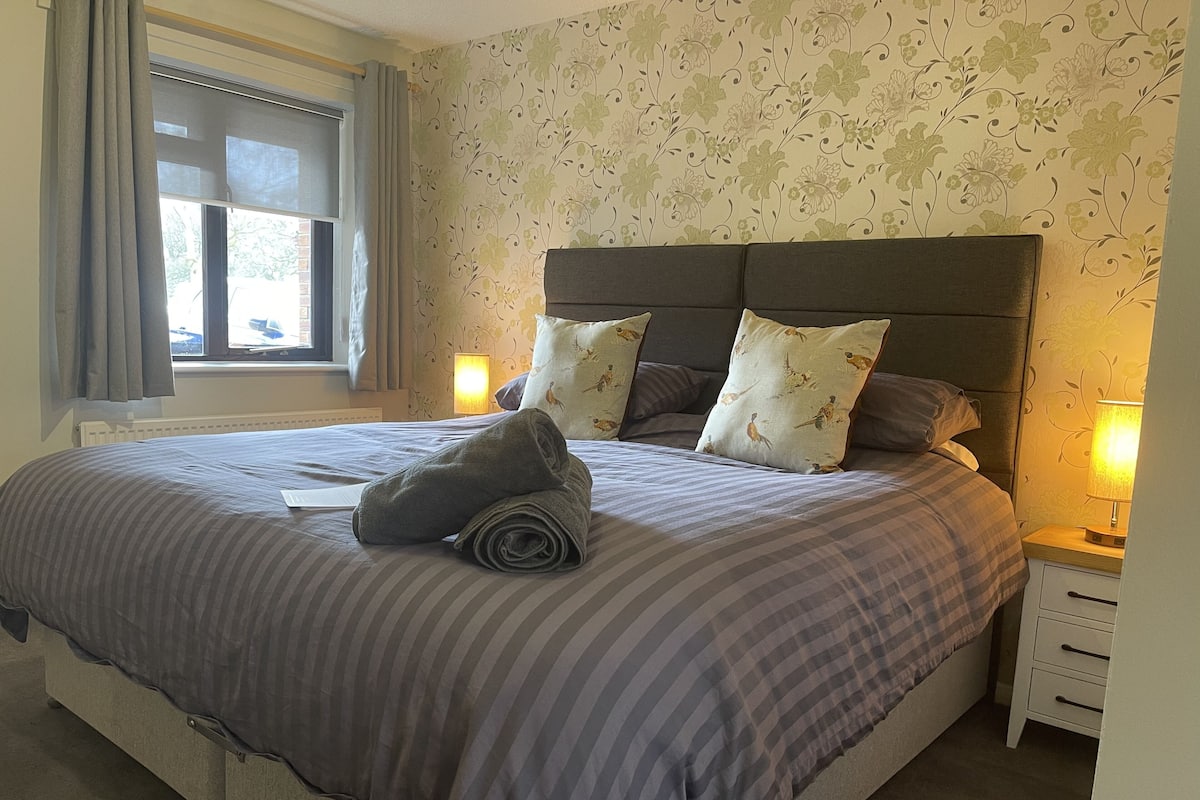 A neatly made bed with striped bedding, two pillows with a bird pattern, and a rolled towel on the bed. There is a window with curtains, a nightstand with a lamp, and a wall with a floral wallpaper pattern.