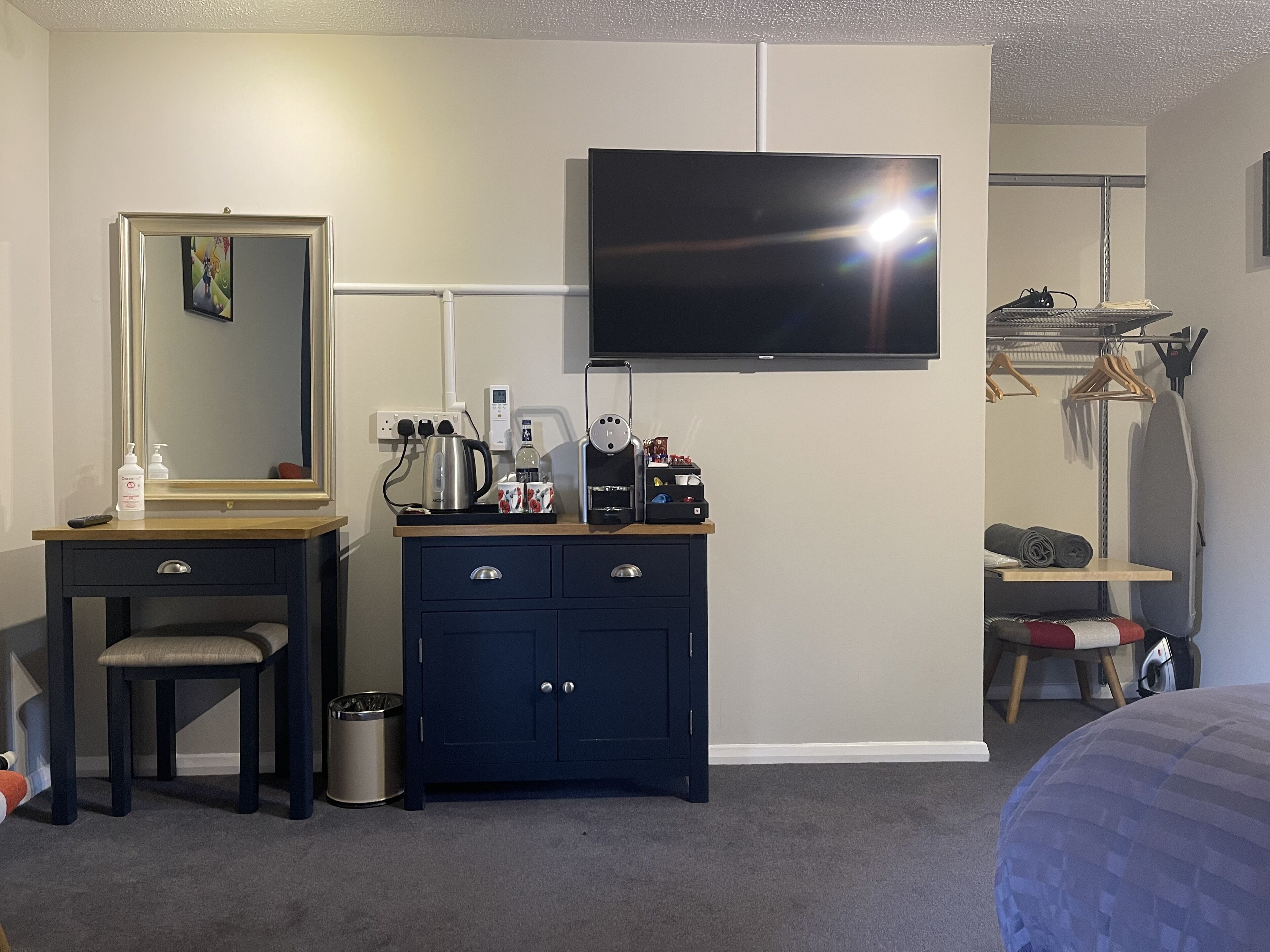 A hotel room with a bed, a desk with a chair, a dresser, a TV mounted on the wall, a mirror, and a small shelf with toiletries.