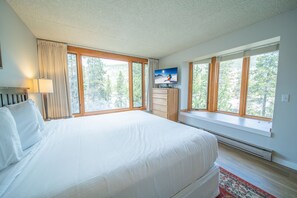 Un dormitorio con una cama grande, un televisor en la pared y una ventana con vistas a árboles.