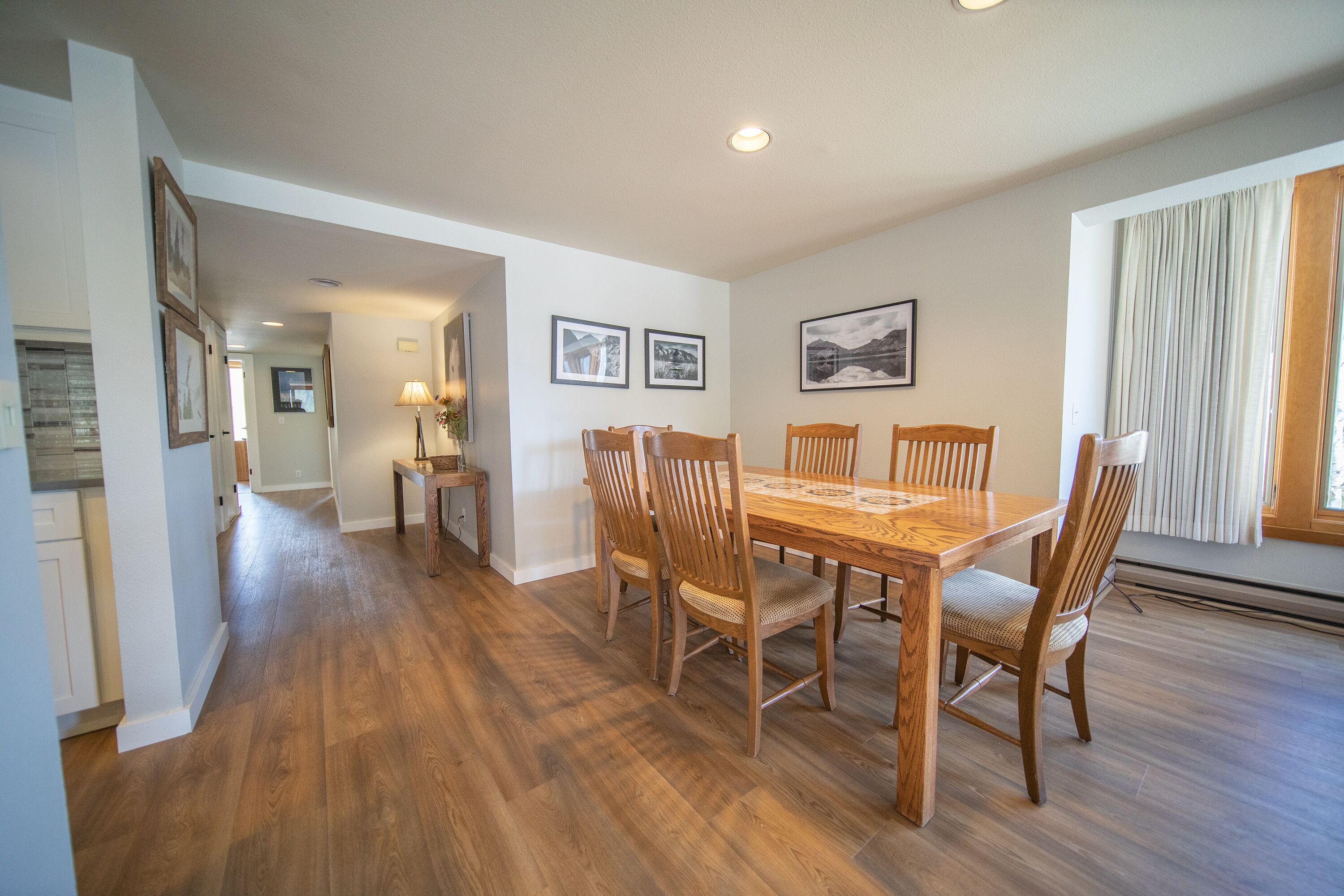 A dining area with a wooden table and chairs, framed pictures on the wall, and a window with curtains.