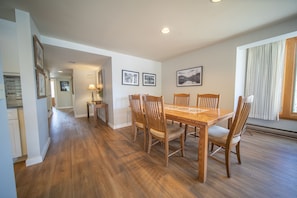 A dining area with a wooden table and chairs, framed pictures on the wall, and a window with curtains.