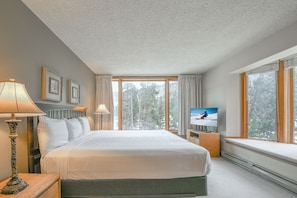 A hotel room with a large bed, a television, a window with a view of trees, and a lamp on the side table.