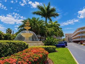 A fountain with multiple tiers, surrounded by well-maintained hedges and flowering plants, with a clear blue sky and a building in the background.