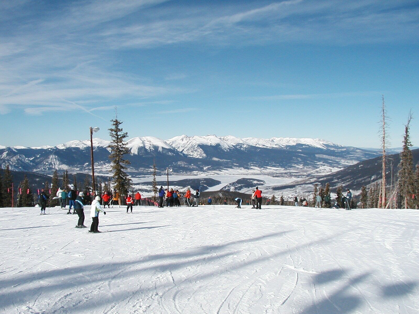 A ski slope with skiers and snowboarders, snow-covered mountains in the background, and a clear sky.