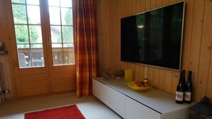 A room with a wooden paneled wall, a large flat-screen TV mounted above a white console table, a red rug on the floor, and a window with curtains. - Halten (Hofmann) by Interhome (Zweisimmen)
