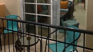 En balkon med turkis farvede stole, et glasbord og en skydedør, der fører til en opholdsstue med en sofa og et fjernsyn.
