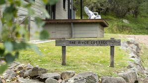 A wooden sign with the text "THE RIVER COTTAGE" in front of a building with a porch and a chair. - The River Cottage - Idyllic river side cottage (Joadja)