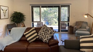 A cozy living room with a brown leather sofa, patterned cushions, a side table, a floor lamp, and a large window with a view of trees. - The River Cottage - Idyllic river side cottage (Joadja)